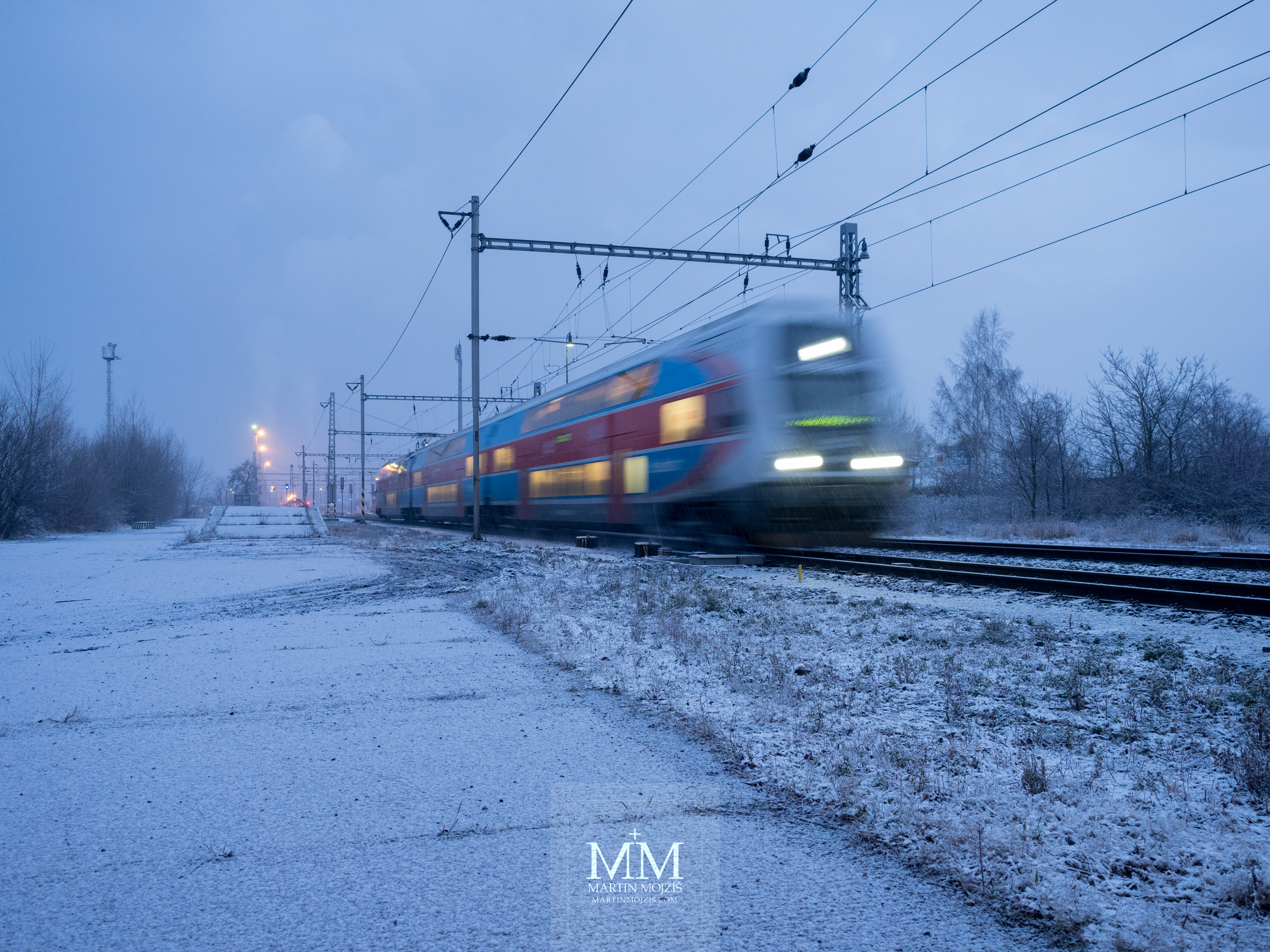Vlak odjíždí ze zasněžené stanice v mrazivém zimním ránu. Fotografie vytvořena objektivem Olympus 12 - 40 mm 2.8 Pro.